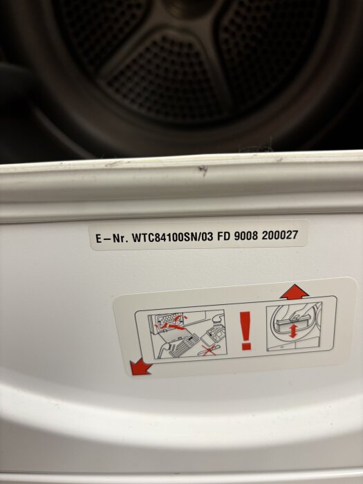 Label med serienummer och varningar på vit yta inuti vitvarumaskin, med instruktioner för borttagning eller justering.