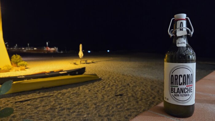 Flaska med Arcana Blanche öl på ett bord nära en strand under natten.