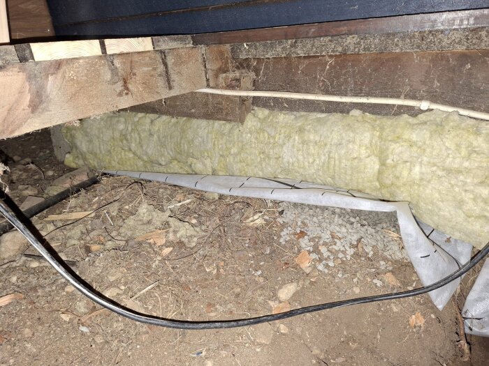 Isolering lagd direkt på marken under golvet i trästuga, utan luftgap, med synlig kabel och byggmaterial i krypgrund.