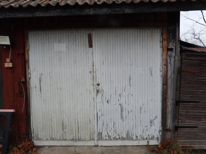 Sliten vit garageport med rostiga handtag, slitna brädor och sprucken färg, omgiven av röd träpanel och trätak med tegelpannor.