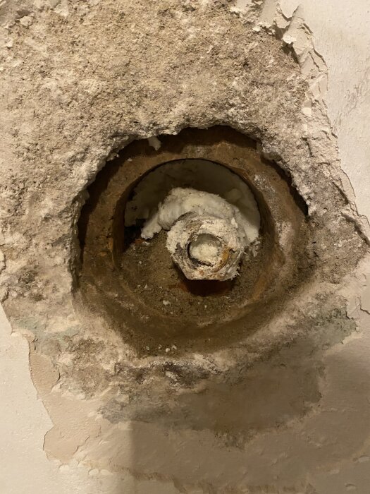 Gammalt igensatt rör i källarvägg, fyllt med fogskum och stenar, del av vattenledning. Synlig slitage på väggens puts runt röret.