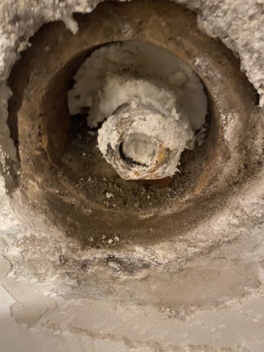 Gammalt pluggat rör i källarvägg, fyllt med sten och fogskum; synliga spår av puts och fukt runt rörets kant.