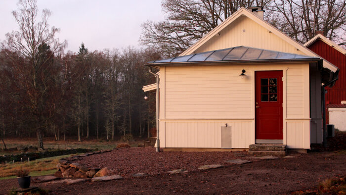 Vit sidobyggnad med röda dörrar och plåttak på grusad mark i skogsområdet.