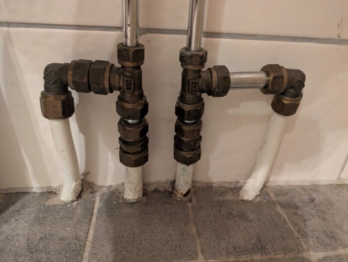 Rörkopplingar i ett badrum med två vita rör som kommer upp från golvet och två metallrör som går in i väggen, del av ett golvvärmesystem.