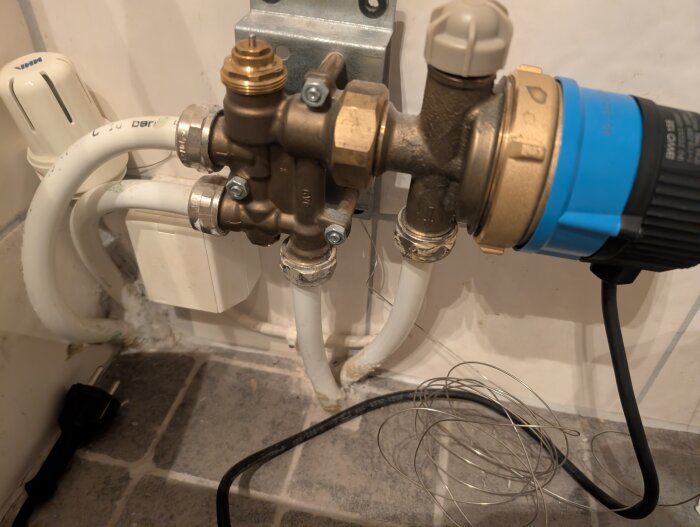 Vattenrör och kopplingar till en cirkulationspump för golvvärme syns i ett badrum med synliga kablar.