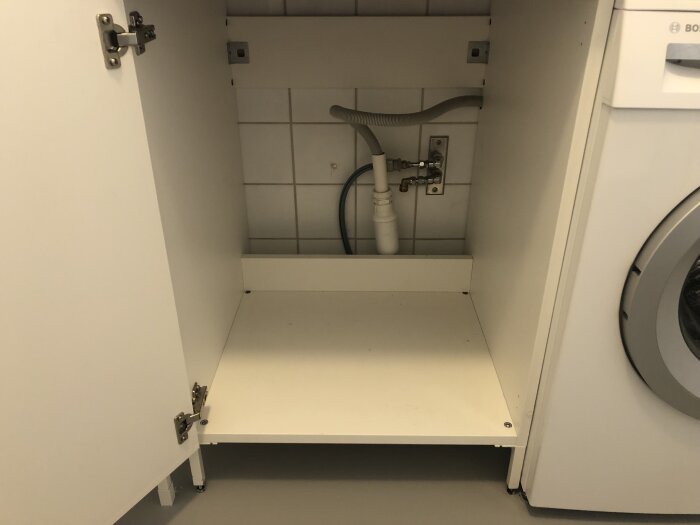 Skåp under bänkskiva med rör och ventiler synliga mot en vit kakelvägg. Vänsterdörren är öppen och till höger syns en del av en tvättmaskin.