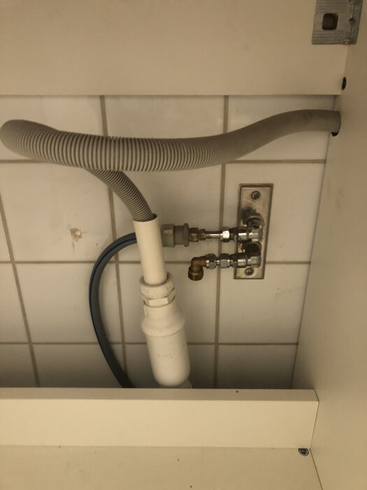 Vattenrör och slangar under bänkskivan i ett badrumsskåp, med väggplattor i bakgrunden.
