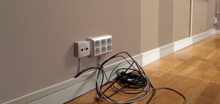 Elektriska uttag och kablar längs väggen ovanpå ett trägolv, med en kanal för kabeldragning.