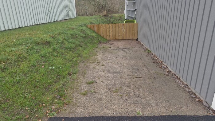 Gräsbevuxen slänt bredvid grått garage med enkel trävägg i slutet av en grusbelagd yta. Gula verktyg syns i bakgrunden.