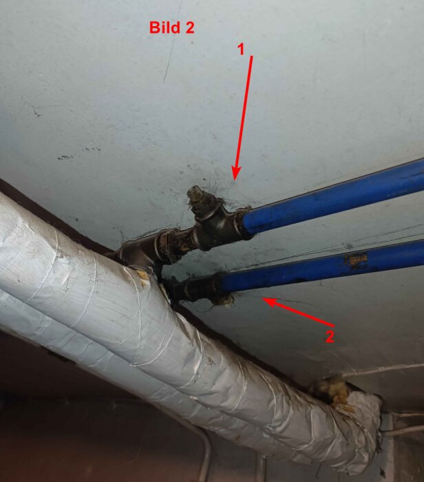 Två blå rör anslutna till ett T-rör. Det övre röret (1) går in i T-röret, och det undre röret (2) ansluts till en större ledning täckt av isolering.