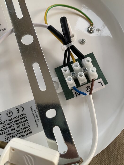 Elektrisk kopplingsplatta med kablar anslutna, en gul-grön jordkabel och en blå och brun kabel. Breddad metallvinkel syns också.