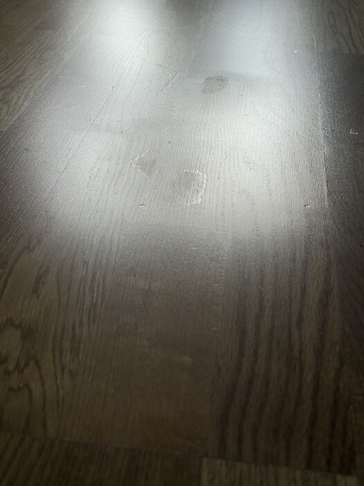 Små blanka fläckar på ekparkettgolv syns vid släpljus, troligen från borttagning av tejprester som dragit med lacken.