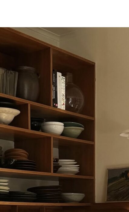 Trähylla med olika keramikskålar, tallrikar, kannor och böcker arrangerade i fack mot en beige vägg.