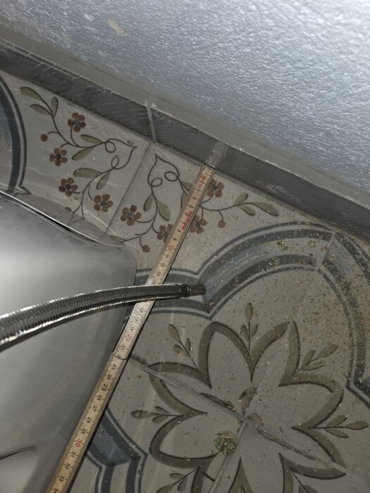 Mätsticka ligger på klinkergolv vid vägg, nära ett rör och en toalettstol. Golvet har blomliknande mönster och väggen är kaklad.