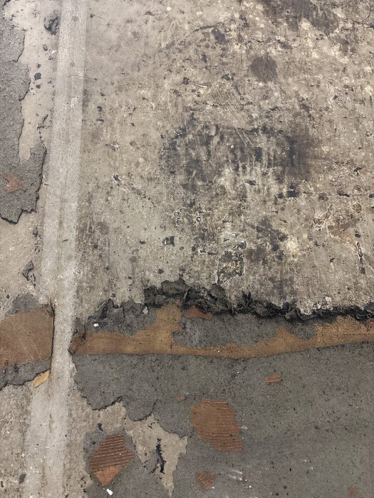 Betonggolv med rester av lim och gammal matta, delvis avskrapat. Ytan är ojämn och visar slitage.