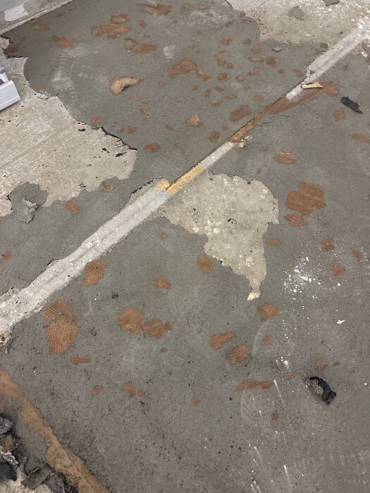 Betonggolv med rester av lim och gammal matta som ska tas bort, delar av betongen är blottade och fläckar av brunt lim syns spridda över ytan.