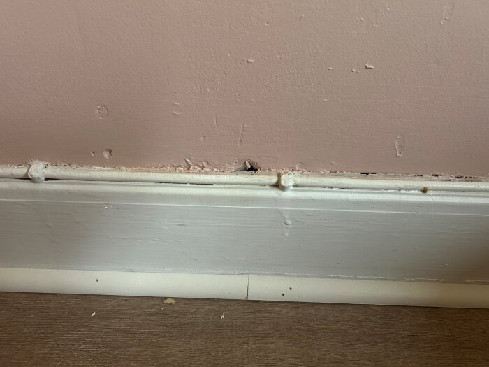 Hålighet mellan vägg och golvlist med synligt drag i ett rum, sprickor i rosa vägg ovanför vit list, förslag på tätning med akrylfog.