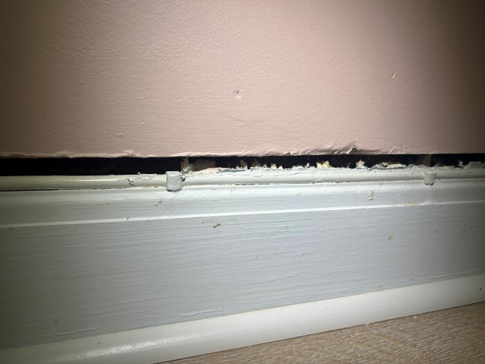 Hål i väggen ovanför en vit golvlist, med synliga mellanrum och drag, på en rosa vägg där fogning och tätning efterfrågas.