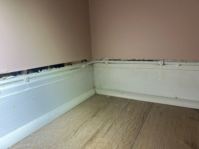 Hål i väggen mellan golvlist och rosa vägg i ett hörn av rummet, med synlig dragproblematik och behov av tätning för att minska värmeförlust.