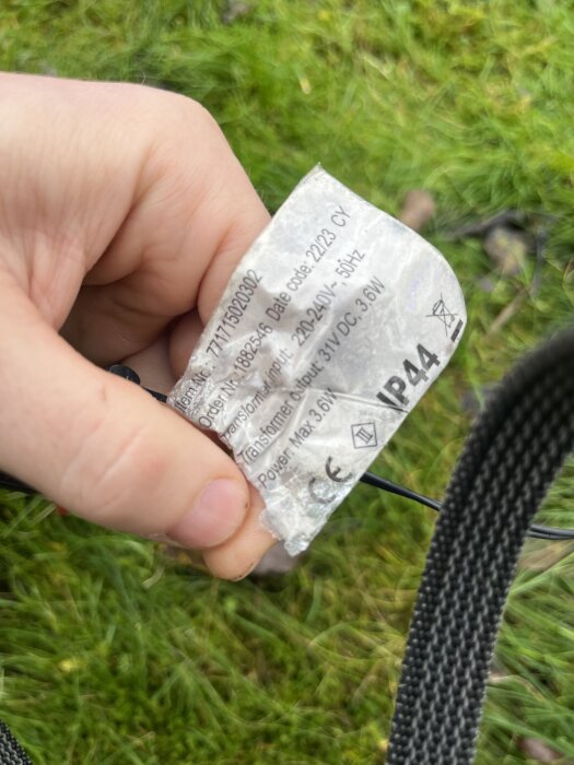Närbild på hand som håller etikett från transformator med specifikationer för LED-slinga på gräs.