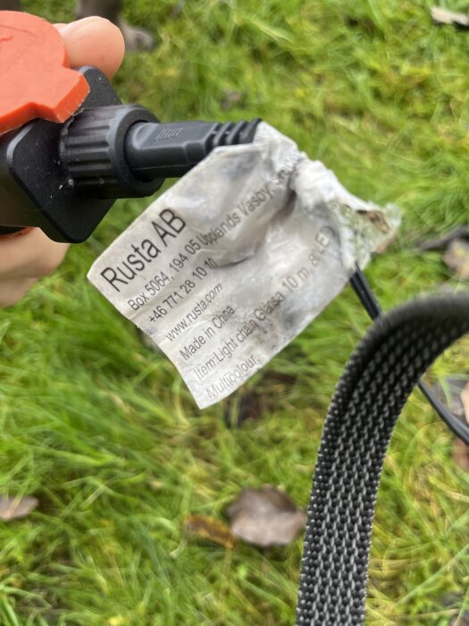 Transformator från Rusta med etikett på gräsmatta, visar modell- och kontaktinformation för ljusslinga, samt sladdsammansättning.