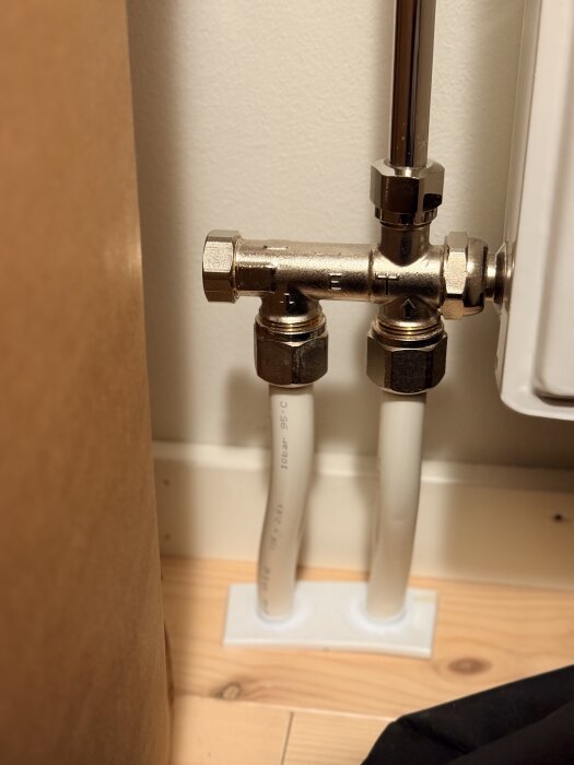 Vita rör med kopplingar syns vid golvet under ett vatten-element. Röret till vänster är märkt med tryckklass och temperatur.