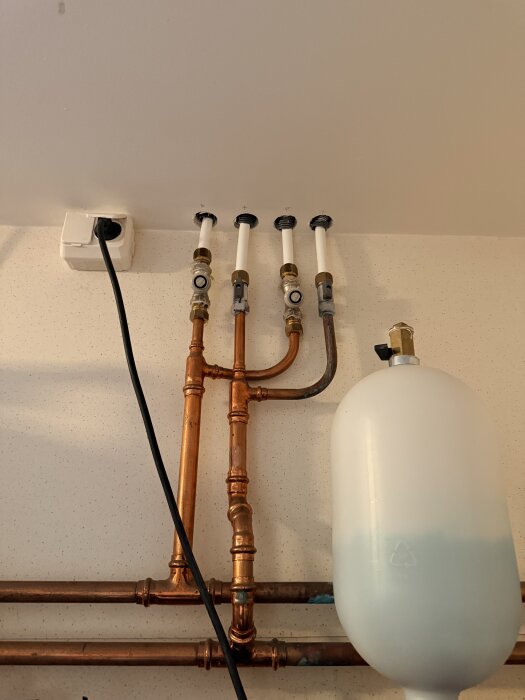 Kopparrör och röranslutningar vid taket med fokus på rör som leder till element, samt en vit expansionstank på väggen.