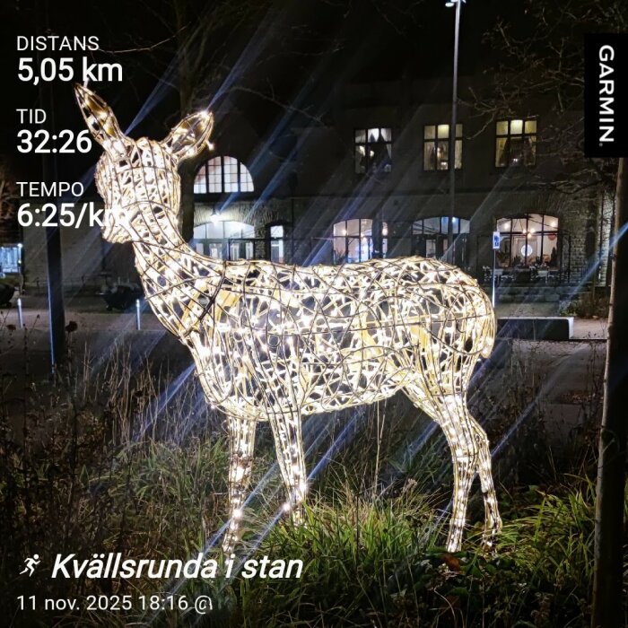 Lysande hjortskulptur av metall och ljus på en gräsmatta, omgiven av mörker och bakgrund av byggnader.