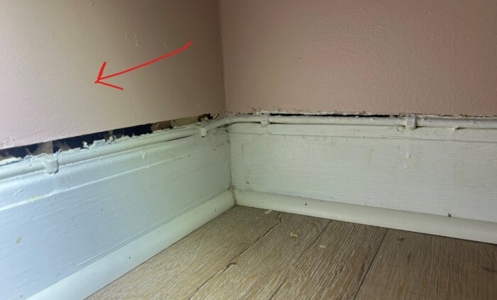 Vägg med rosa färg, bruten trälist vid golv, gapande hål bakom, röd pil pekar på väggen, linoleummatta syns på golvet i hörnet.