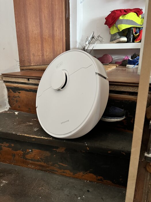 Vit Dreame robotdammsugare på en trappa framför en garderob fylld med skor och kläder i olika färger.