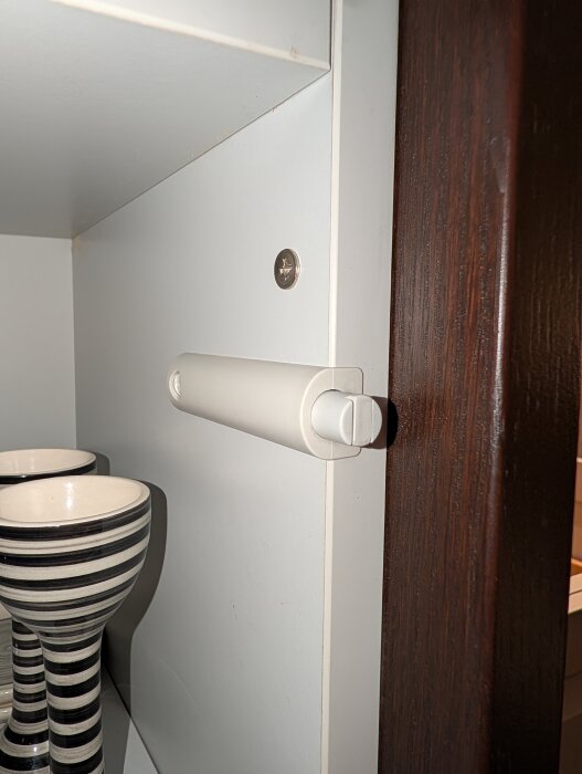 Push-to-open mekanism på köksskåpsdörr, vit och cylindrisk, med två randiga muggar i bakgrunden.