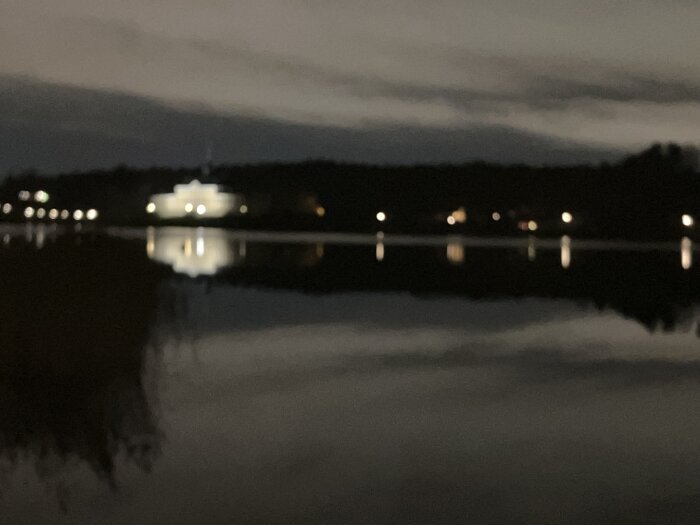 Osäker bild av ett naturskönt landskap i skymningen med en byggnad och ljusreflektioner i vattnet.