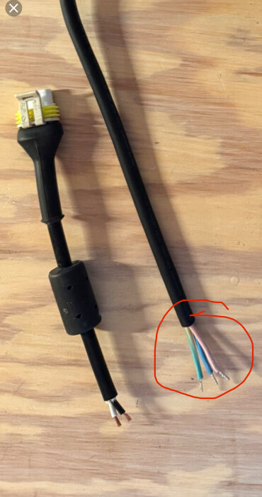 Två elektriska kablar på träyta, den ena med flerfärgade ledare exponerade och markerade med röd cirkel.