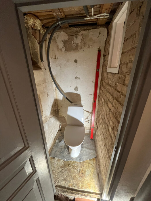 Toalett i källare under konstruktion; osäker golvyta med grå matta, röd vattenpass lutad mot väggen, ledningar synliga i taket.