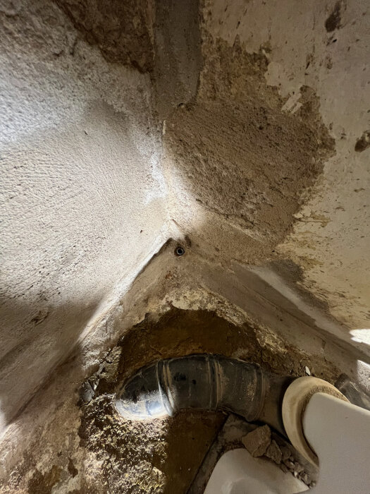 Gammal gjutjärnsstam och golv i källare. Väggen och hörnfogar är delvis skadade och golvet har brutits upp runt röret för avloppsrenovering.