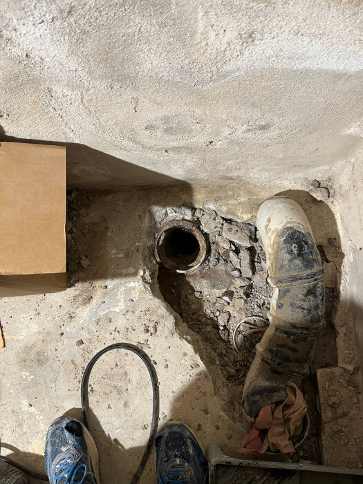 Uppbilat golv med synligt gammalt avloppsrör och betongskada i källaren, sedd ovanifrån. Två blå sneakers syns i bildens nederkant.