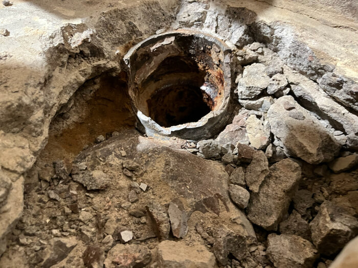 Gammalt gjutjärnsrör i en uppbruten betonggolv med stenar runtom, för ett avloppsprojekt i en källarombyggnad.