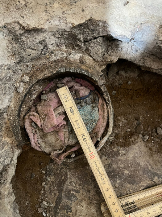 Öppen golvyta med gammalt avloppsrör fyllt med trasor och en mätsticka ovanpå, omgiven av rå betong och jord.