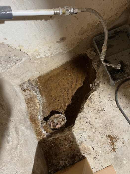 Ett uppbilat hål i källargolvet med synlig rörstump, vit vägg och kopplad slang ovanför.