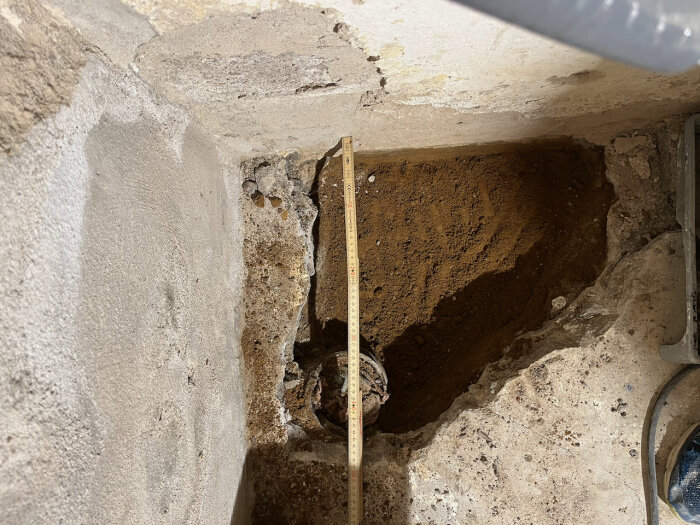 Uppborrat golv med synligt gammalt avlopp i källare, exponerad jord och ett måttband som mäter djupet av hålet.
