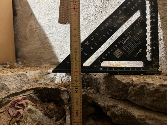 Mätverktyg och tumstock mäter ett uppbilat betonggolv i en källare, omgivna av grus och betongbitar.