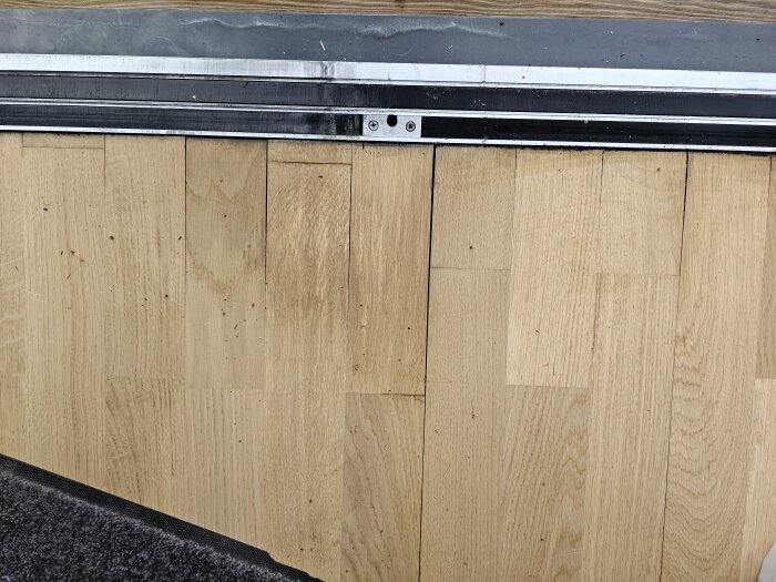 Trägolv med springa där fukt kan tränga in, nära dörrtröskel. Indikation på potentiell vattenskada eller byggproblem.