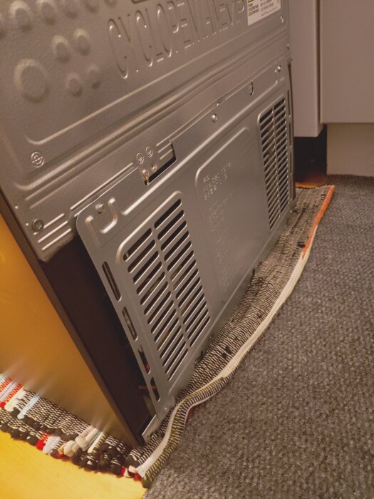 Baksidan av en Samsung-frys utan skyddsplåt; ett kopparrör sticker ut, stående på en matta.