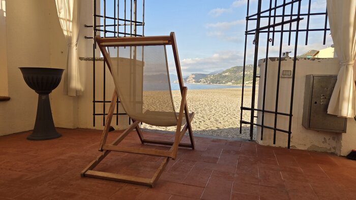 Solstol på en terrass med utsikt över en sandstrand och hav, omgiven av öppna smidesjärngrindar och vita gardiner.