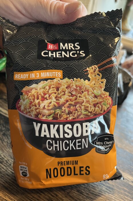 Förpackning med Mrs Cheng's Yakisoba Chicken premiumnudlar redo på 3 minuter, innehåller nudlar och kyckling, hålls i handen.