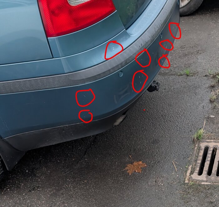 Blå bil bakifrån med flera markerade stenskott på stötfångaren. Aning av grå asfalt och ett galler syns i bakgrunden.