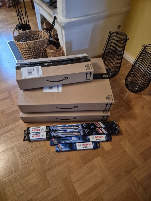 Tre stora Amazon-kartonger med fyra Bosch torkarbladspaket på golvet framför en kamin med vedkorg.