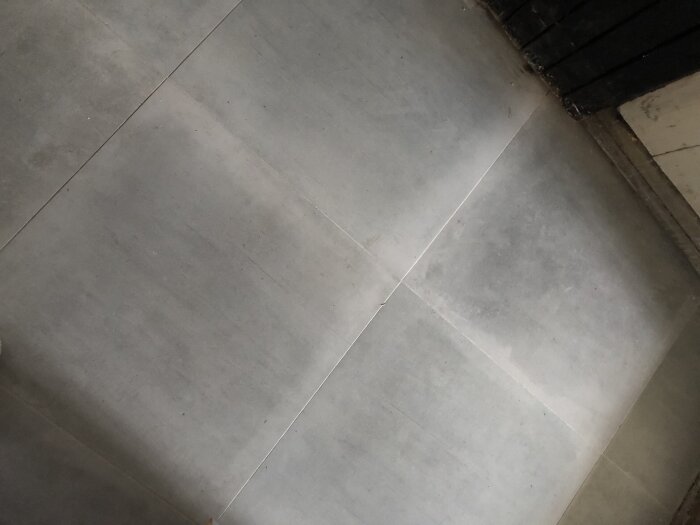 Grått kakelgolv med svarta hörnlister, skimrande yta och synliga fogar.