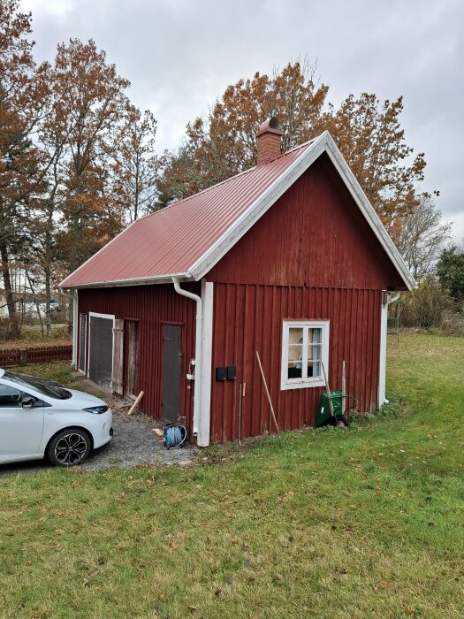 Rött uthus med rött tak och skorsten, omgiven av träd och gräs, med en vit bil parkerad framför.
