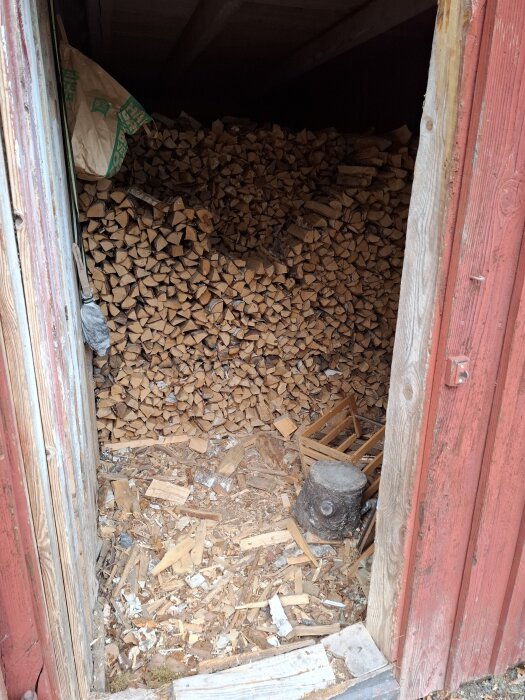 Inuti ett uthus med staplad ved och en stubbe på golvet, omgiven av träväggar.